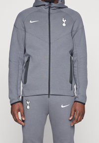 Felpa grigia con zip e cappuccio, logo Nike bianco, emblema degli Spurs, due tasche laterali con zip, polsini a coste e tessuto testurizzato.