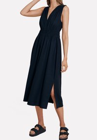 Robe sans manches bleu marine avec un col en V profond, une taille smockée et une fente latérale. Tissu léger avec une silhouette fluide. Portée avec des sandales à plateforme.