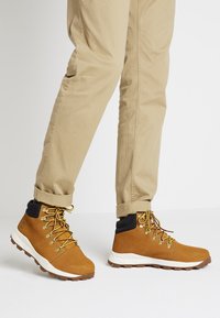 Bruna lädervandringsskor med gula snören, vit sula och svart vristkrage. Bärs med beige uppvikta byxor.