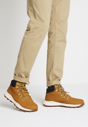 Stivali da trekking in pelle marrone con lacci gialli, suola bianca e colletto nero. Indossati con pantaloni beige arrotolati.