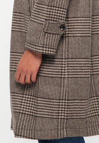 Pruunist ruudulisest kangast tehtud mantel, millel on tekstuurne kangas, ühe nupuga mansett ning nähtavad houndstooth ja ruudulised mustrid.