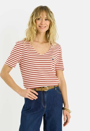 Mujer con cabello rubio y ondulado, vestida con una camiseta de rayas rojas y blancas con escote en V, vaqueros azules, cinturón marrón claro y pulseras doradas, sonriendo con la mano en la cadera.