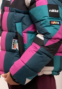 Donsjack met gedurfde geometrische patronen in teal, roze en zwart. Heeft een label en een ritsvak aan de linkerzijde.