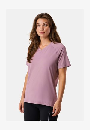 Woman with long brown hair wearing a light purple short-sleeve v-neck shirt and black pants against a white background.