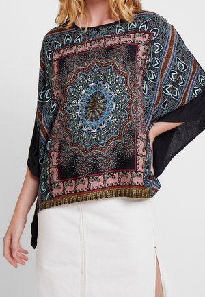 Žena v černém volném topu s barevným zdobeným mandalovým a geometrickým vzorem, kombinovaném s bílou sukní s předním rozparkem.