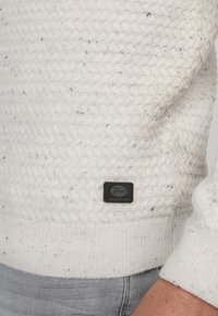 White knitted sweater featuring a textured pattern and a black label. The cuffs and hem are ribbed. Small flecks of color are visible.