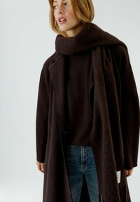 Langer, dunkelbrauner Mantel mit strukturierter Oberfläche, über einem dunklen Pullover getragen und von einem passenden Schal begleitet. Helle Denim-Jeans sichtbar.