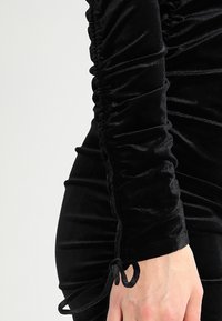 Schwarzes Samtkleid mit gerafften Details an Ärmeln und Seite, ausgestattet mit verstellbaren Kordeln für eine anpassbare Passform. Glatte, weiche Textur.