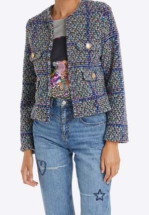 Frau trägt eine mehrfarbige, strukturierte Jacke mit goldenen Knöpfen über einem Grafik-T-Shirt, kombiniert mit hoch taillierten blauen Jeans mit Stern- und Herz-Aufnähern.