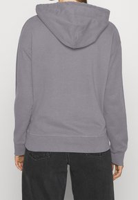 Felpa grigia con cappuccio, dal taglio rilassato, in tessuto testurizzato e con cuciture visibili. Indossata con jeans scuri, che mostrano una vista posteriore.