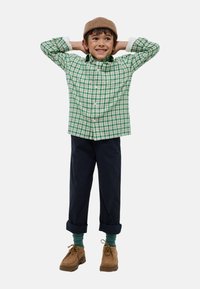 Chemise à carreaux verte et blanche, pantalon bleu marine, chaussures brunes et bonnet en tricot beige. Mains levées, avec des chaussettes vertes visibles.