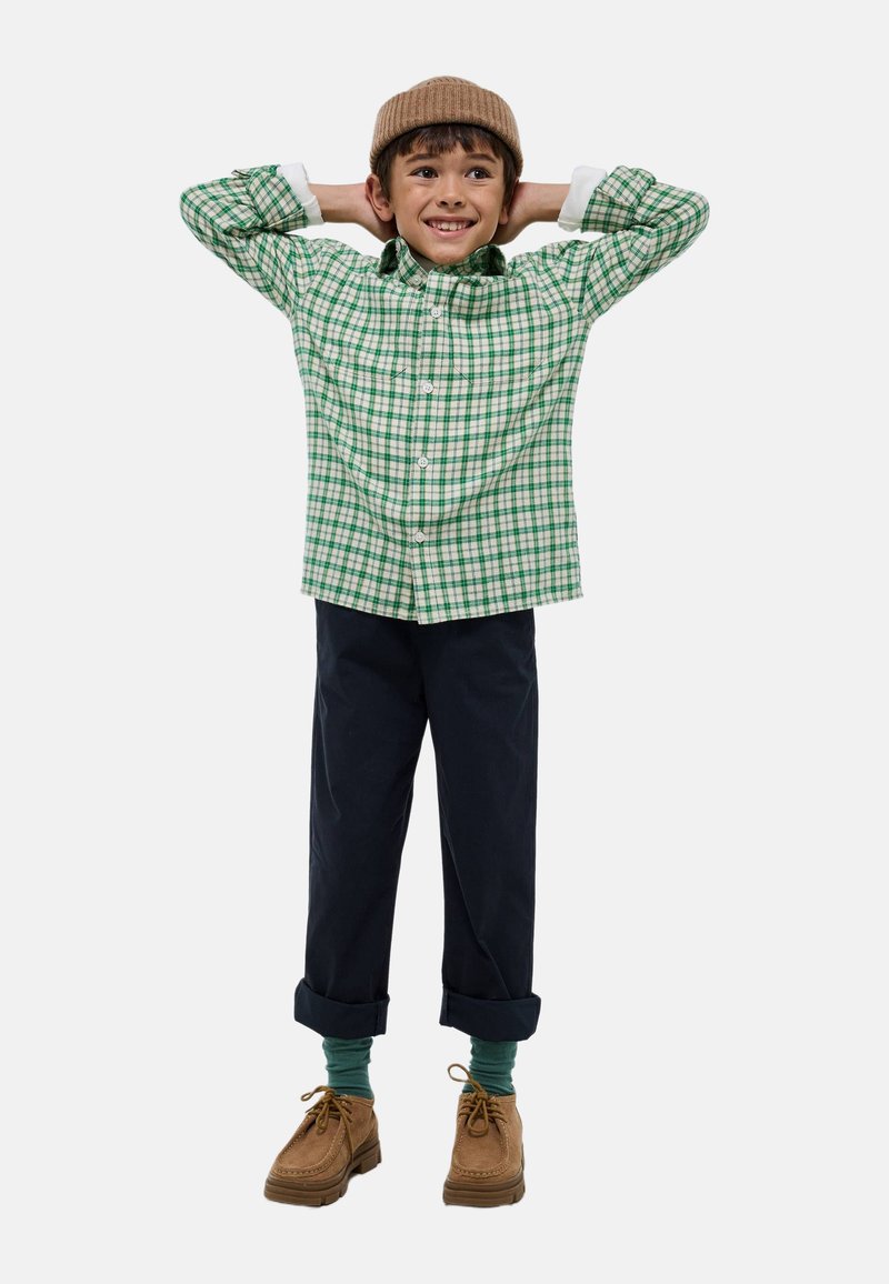 Chemise à carreaux verte et blanche, pantalon bleu marine, chaussures brunes et bonnet en tricot beige. Mains levées, avec des chaussettes vertes visibles.