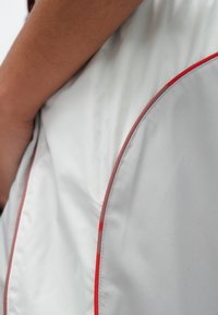 Biała kurtka o gładkiej fakturze z czerwonymi akcentami wzdłuż krawędzi. Zbliżenie ukazuje połysk tkaniny oraz zakrzywione elementy wzornicze.