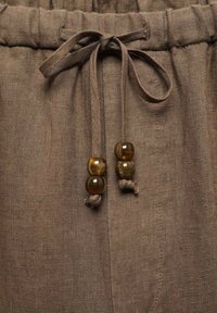 Pantalon en lin marron avec une taille à cordon, comprenant deux perles décoratives marron sur les liens et une surface en tissu texturée.