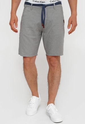 Mand iført grå shorts med nålestriber med blåt bælte, hvid Calvin Klein linning og hvide sneakers, stående mod hvid baggrund.