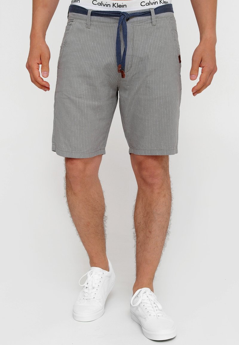Homme portant un short gris à fines rayures avec ceinture bleue, ceinture blanche Calvin Klein et baskets blanches, debout devant un fond blanc.