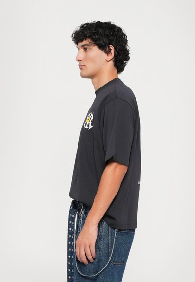 Hombre joven con cabello rizado y oscuro, vistiendo una camiseta gráfica negra y jeans azules con un cinturón de ojales de metal, de perfil frente a un fondo sencillo.