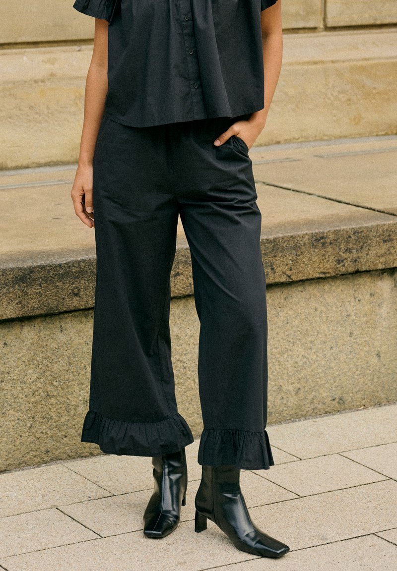 Femme portant un pantalon noir à jambes larges avec des ourlets à volants et des bottines noires à talons, debout sur un pavé en pierre.