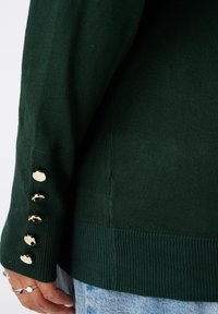 Groene gebreide trui met geribbelde boorden en een zijschuif, versierd met gouden knopen langs de mouw voor extra detail.