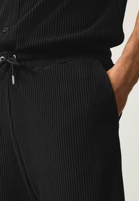 Pantalones negros acanalados con cintura ajustable por cordón, bolsillos laterales y tejido texturizado que resalta líneas verticales.