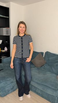 Γυναίκα που χαμογελά, φοράει μπλε-σκούρο και λευκό πουκάμισο με ρίγες και κουμπιά, μπλε τζιν και λευκά αθλητικά παπούτσια, στέκεται δίπλα σε μπλε καναπέ από κοτλέ και μαύρη μονάδα ραφιών.