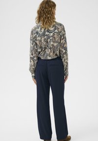 Osoba s kučeravými vlasmi, oblečená v blúzke s paisley vzorom a tmavomodrými nohavicami s širokými nohavicami, stojí so zavretými zádami proti jednoduchému pozadiu.