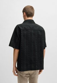 Maglietta nera a maniche corte con design in pizzo floreale, tessuto testurizzato e colletto. La vista posteriore mostra dettagli a motivo e una leggera lucentezza.