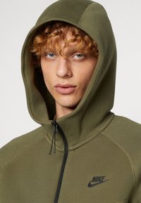 Olive green zip-up hoodie made from soft fabric, featuring a black Nike logo, adjustable drawstring, and a hood with a rounded shape.