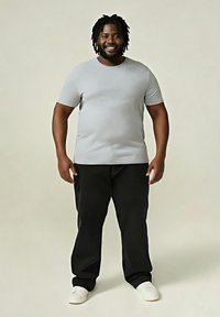 Hombre sonriente con rastas de pie vestido con una camiseta gris clara, pantalones negros y zapatillas blancas sobre un fondo liso.