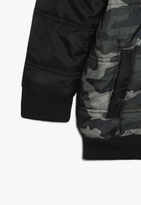 Chaqueta acolchada de camuflaje en negro y gris con puños acanalados y tejido liso contrastante. Cuenta con un bolsillo lateral y textura acolchada.