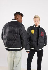 Black puffer jackets with white accent ribbing, featuring colorful embroidered patches: a smiley face and stylized lettering. Lightweight, quilted design.
