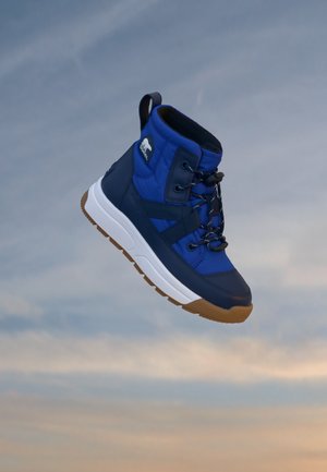 Niebiesko-granatowy sneaker typu high-top z białą podeszwą i brązowym bieżnikiem, unoszący się na tle miękkiego nieba.