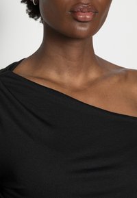 Sort asymmetrisk top lavet af glat stof, med en enkelt off-shoulder design og en draperet tekstur. Subtil glans synlig.