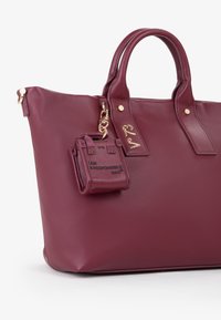 Sac en cuir bordeaux avec double poignée supérieure, accents dorés et un porte-clés mini détachable arborant le texte "JE SUIS UN SAC RESPONSABLE."