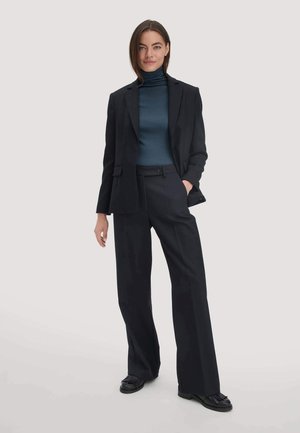 Zwarte op maat gemaakte blazer met piekflappen, gecombineerd met hoge zwarte broeken. Het model draagt een donkerblauwe coltrui. Getextureerde zwarte schoenen.
