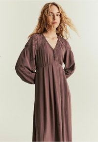 Długa, zwiewna sukienka w przykurzonym mauve z głębokim dekoltem w kształcie litery V, haftowanymi kwiatowymi detalami i luźnymi rękawami, uszyta z miękkiego materiału.