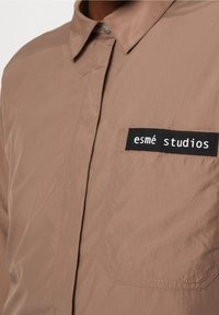 Detailní pohled na hnědou košili s límcem, náprsní kapsou a černou etiketou s nápisem "esmè studios" vyšitý nad kapsou.