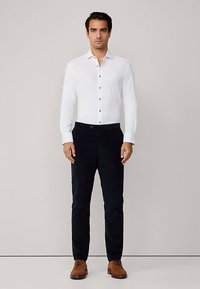 Weißes Hemd mit geradem Kragen und braunen Knöpfen, kombiniert mit dunkelblauen Hosen und braunen Schuhen. Glatte Stoffstruktur.