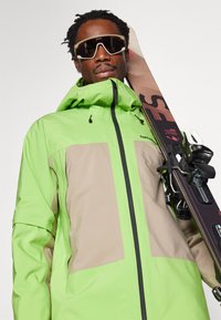 Chaqueta de esquí verde y beige con cierre zip, capucha y bolsillos de ventilación. Gafas de sol oscuras y esquís sostenidos al hombro, mostrando un diseño elegante.