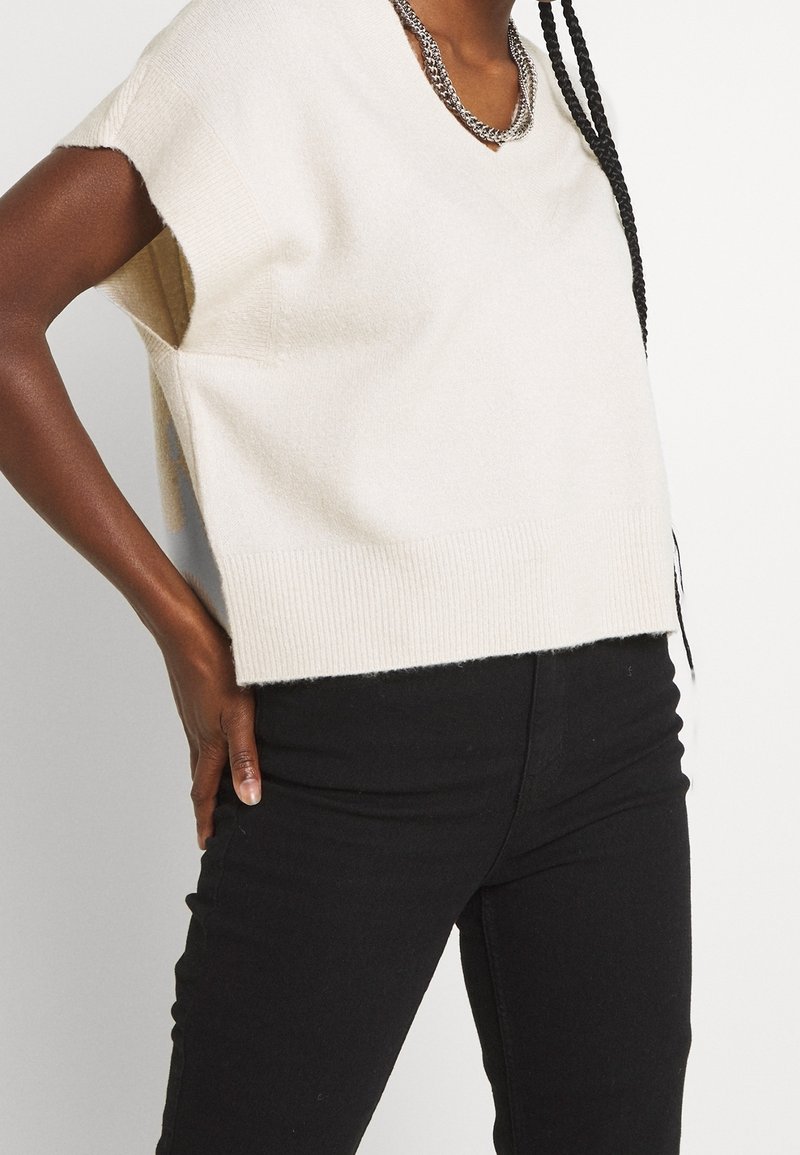 Femme portant un haut en maille crème à manches courtes, pantalon noir, collier chaîne argenté, avec de longs cheveux tressés tombant sur l'épaule.