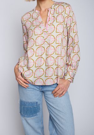 Femme portant un chemisier à motifs avec des formes géométriques roses et dorées, associé à un jean bleu clair avec un grand patch en denim sur le genou.