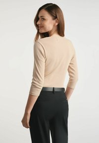 Kobieta z brązowymi włosami ubrana w beżową bluzkę z trzema kwartami rękawów i czarne spodnie, spoglądająca przez ramię na tle jednolitego białego tła.