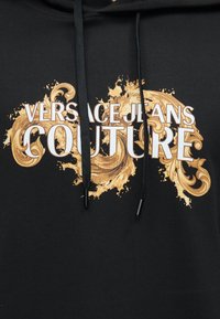Μαύρο φούτερ με χρυσά διακοσμητικά στροβιλίσματα και λευκό έντονο κείμενο "Versace Jeans Couture" στο στήθος, με μαύρα κορδόνια με μεταλλικές άκρες.