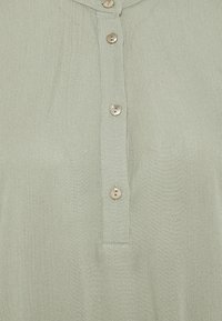 Blusa verde chiaro con apertura a bottoni frontale, cinque bottoni metallici rotondi, una texture sottile e un colletto.
