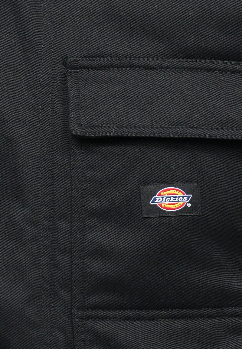 Primo piano di un tessuto nero con una pattina della tasca cucita e un piccolo patch rettangolare del marchio Dickies cucito sulla tasca.