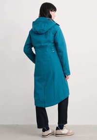 Parka impermeabile verde acqua con cappuccio, bottoni a pressione, due tasche laterali e una silhouette lunga, abbinata a jeans scuri e scarpe beige.