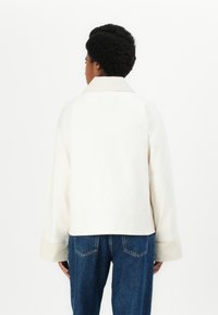 White cropped jacket with a wide collar and loose sleeves, made from a smooth fabric. Paired with blue denim jeans featuring back pockets.