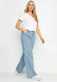 Vaqueros de pernera ancha azul claro con bolsillos frontales, cintura alta y costuras en contraste, combinados con una camiseta blanca de cuello en V y zapatillas blancas.
