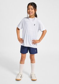 Vit kortärmad sporttröja med svart logotyp, kombinerat med marinblå shorts. Vita knästrumpor och beigea sportskor fulländar looken.