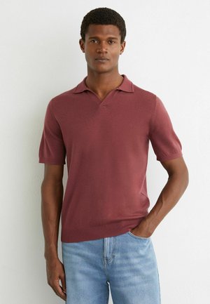 Mees seisab valge seina vastu, kandes lühikeste varrukatega burgundiapunast polo särk ja helehalli teksapükse, üks käsi taskus.